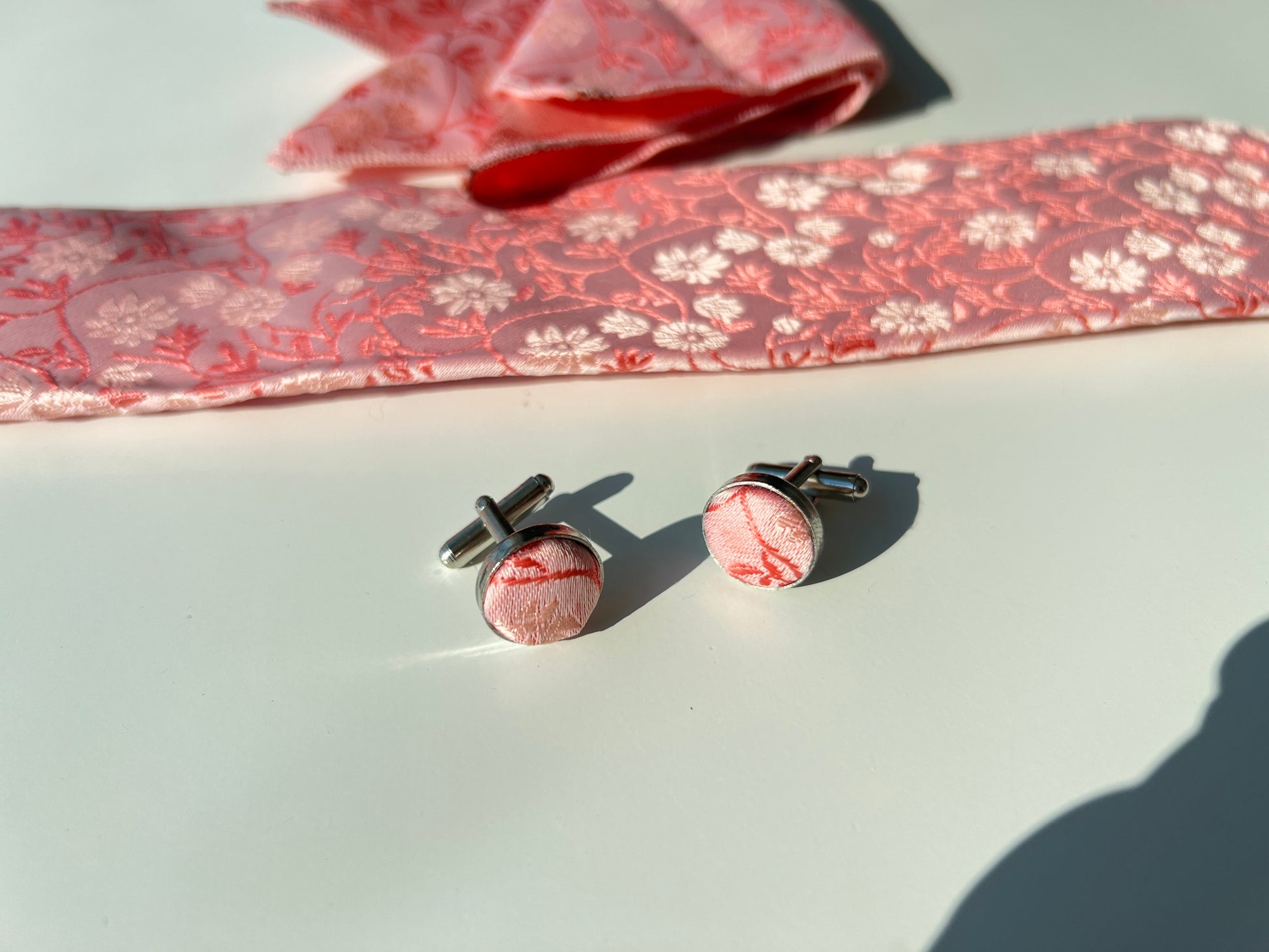 Close-up view of floral pink silk tie, pocket square, and cufflinks showcasing intricate woven details and textures against a sunlit backdrop