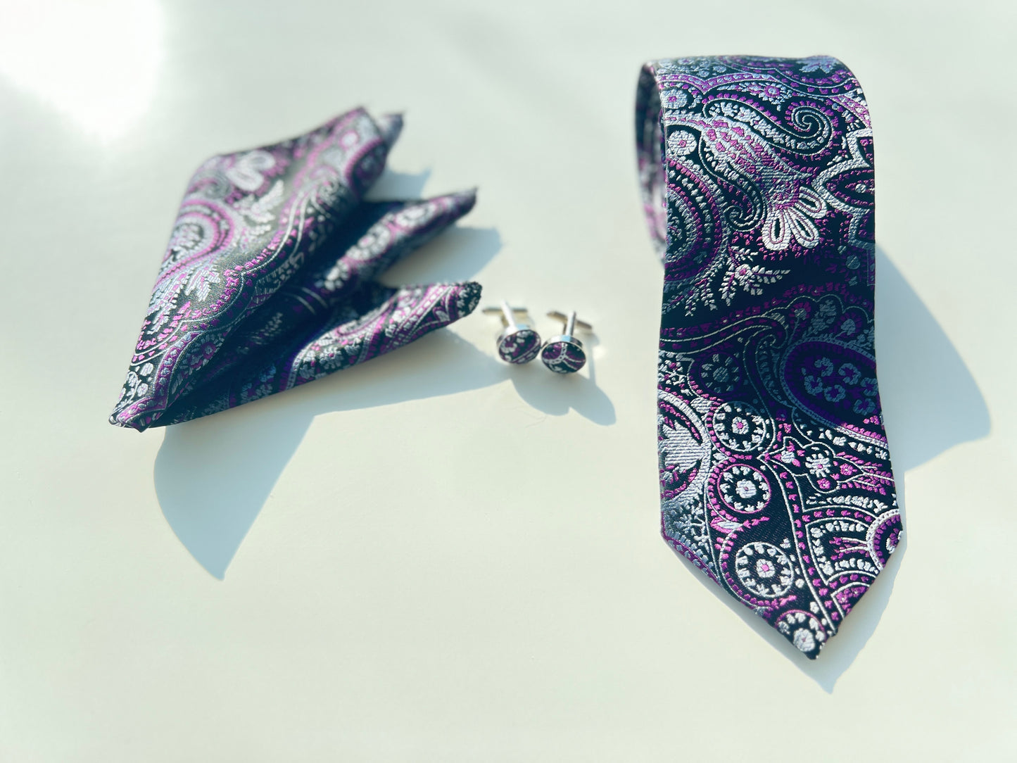 Detailed shot of a luxurious purple paisley tie and cufflinks, with a pocket square, on a sunny day, showcasing the tie’s detailed patterns and vibrant color palette