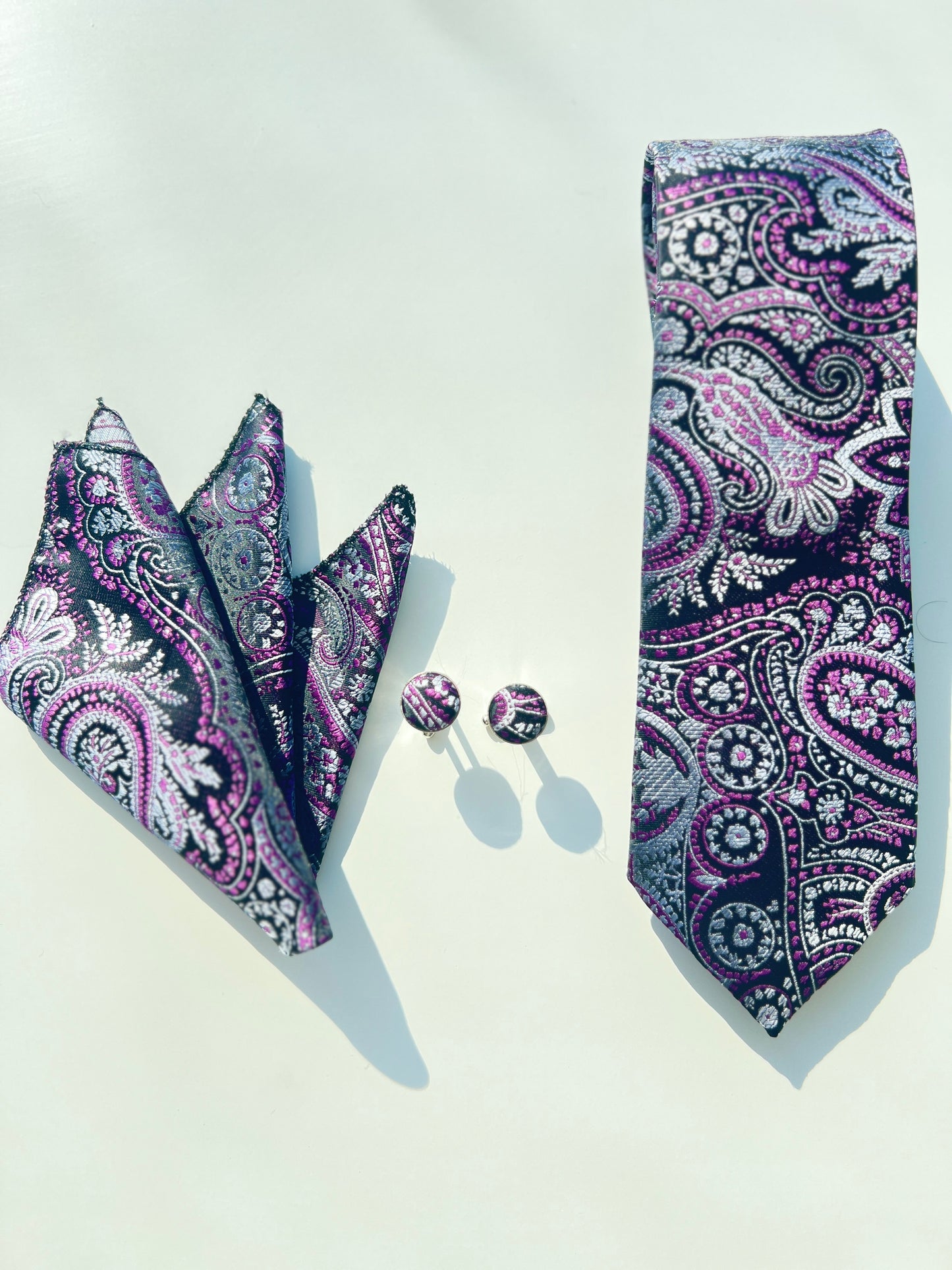 Elegant purple paisley tie set with matching cufflinks and a folded pocket square, displayed on a bright white background, highlighting intricate silver and pink floral designs