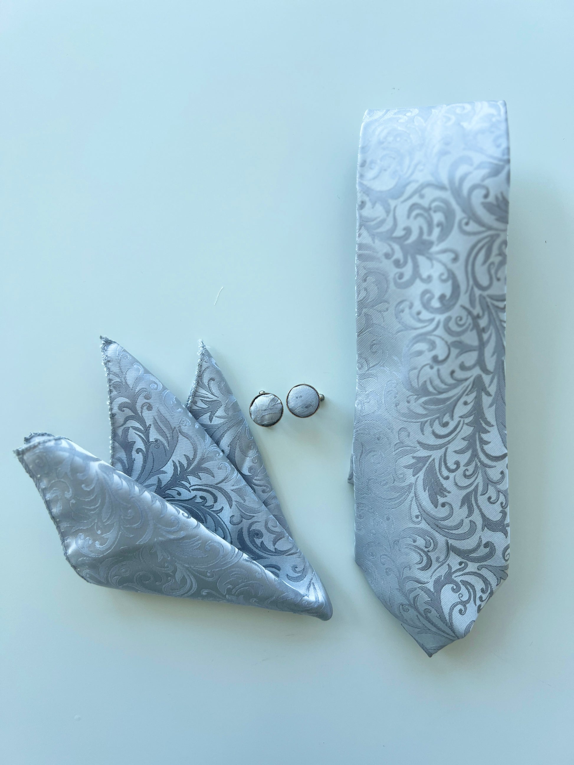 Luxury Silver Paisley Tie Set displayed on a white surface, including a silk tie, pocket square, and pair of cufflinks under natural light