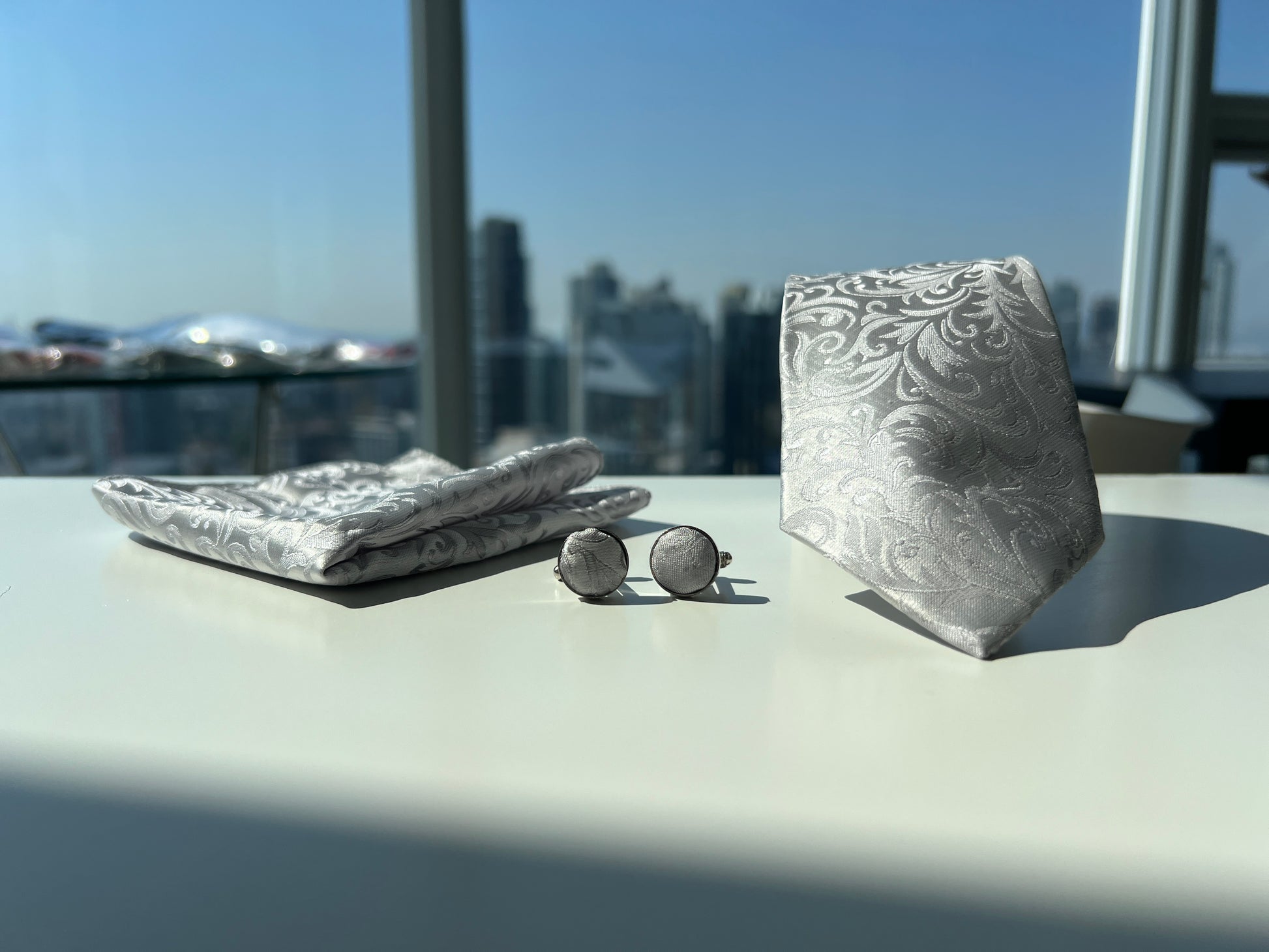 Full view of the silver paisley tie set on a white table, showcasing the tie, pocket square, and cufflinks with a panoramic view of the city skyline