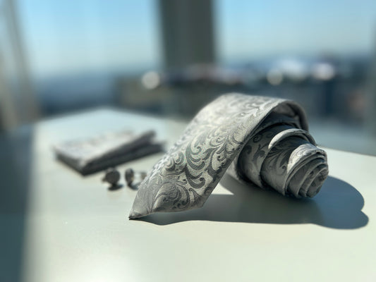 Elegant silver paisley tie partially rolled next to matching pocket square and cufflinks on a white surface, with a high-rise city background