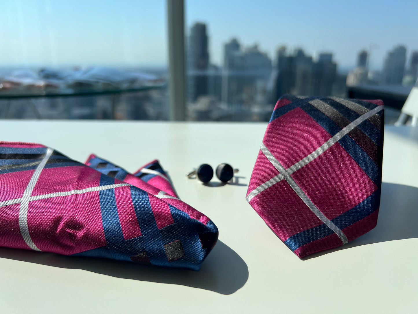 Elegant presentation of the pink and navy striped tie rolled next to matching cufflinks and pocket square, set against a cityscape.