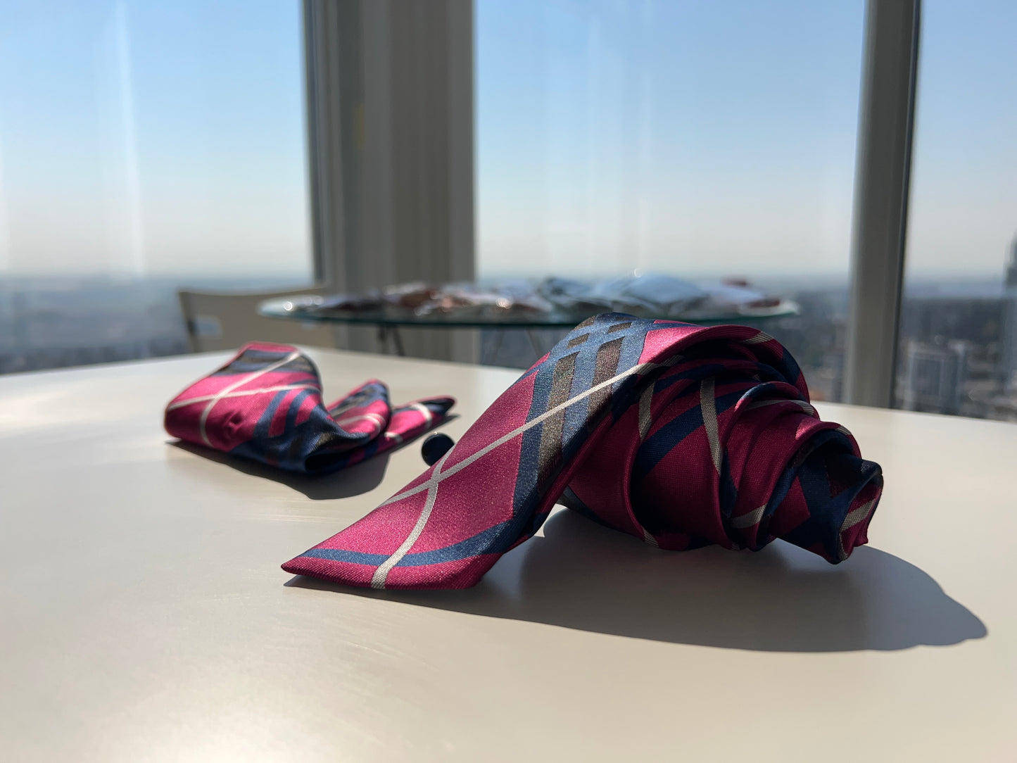 Elegant presentation of the pink and navy striped tie rolled next to matching cufflinks and pocket square, set against a cityscape.