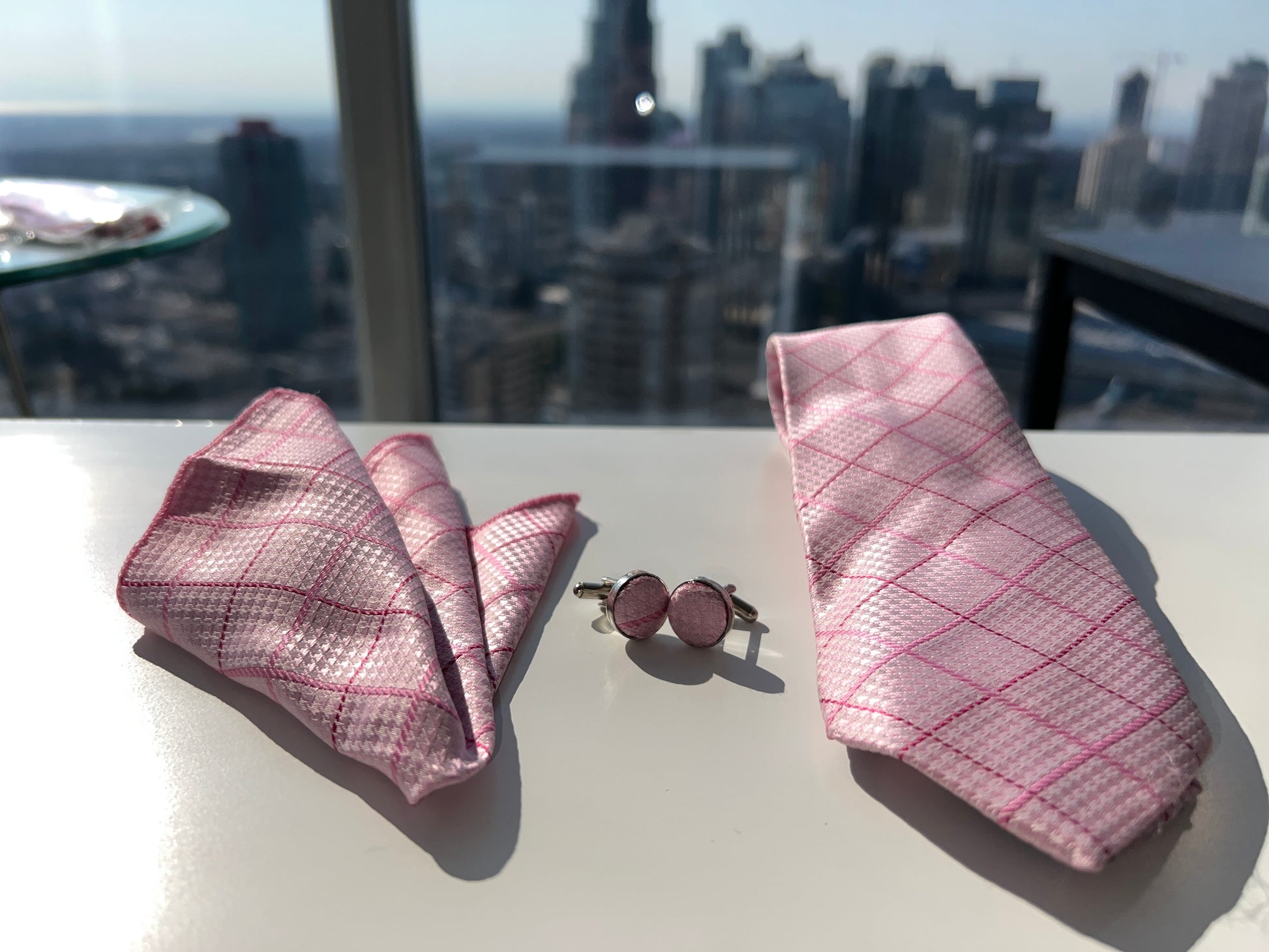 Sophisticated pink tie set with cufflinks, pocket square, presented against a bright background with urban skyline in the distance.