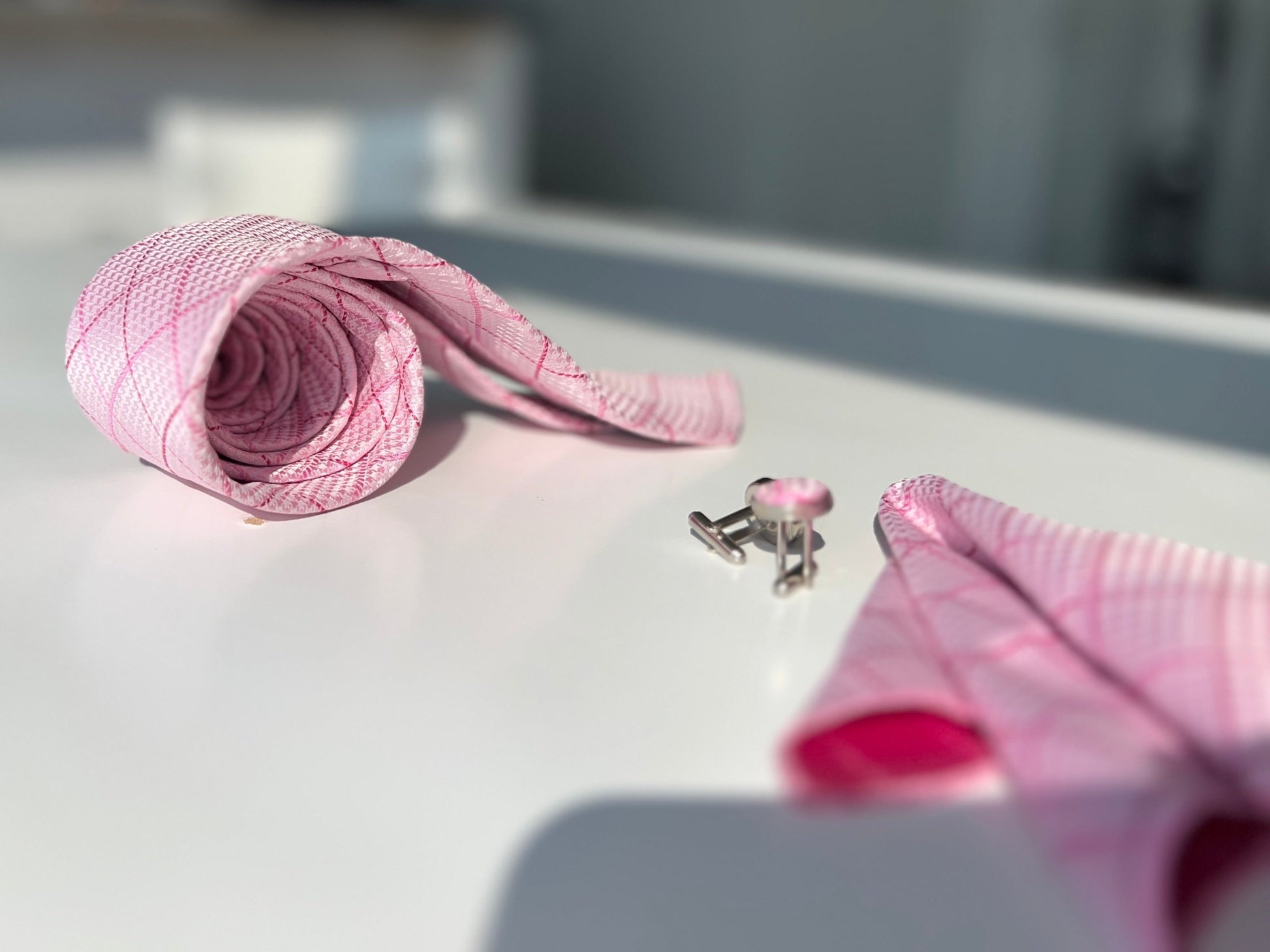 Luxurious pink tie rolled next to cufflinks and a folded pocket square on a white glossy surface, with soft lighting