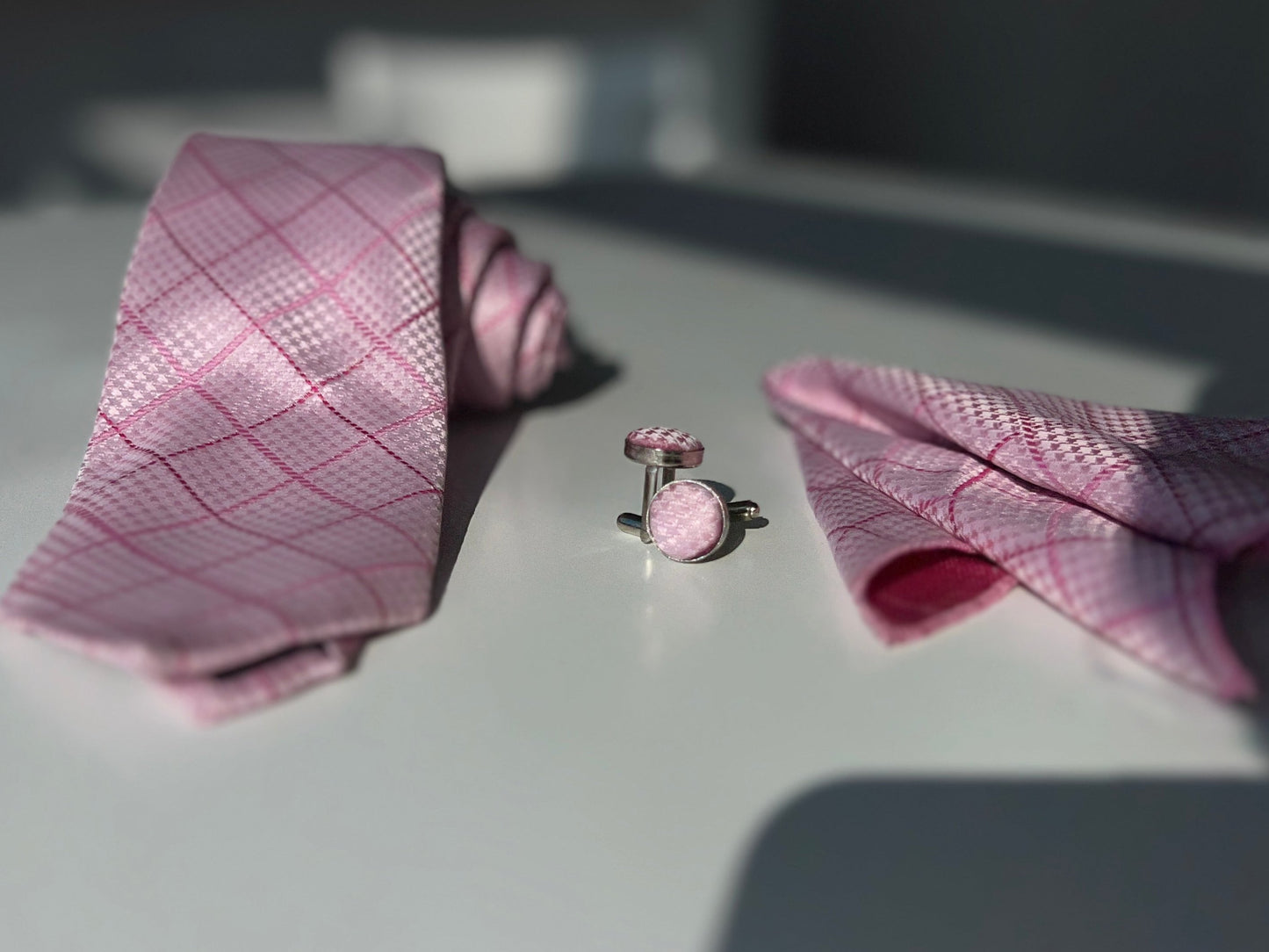 "Close-up of a pink tie set including a tie, pocket square, and cufflinks with a diamond pattern, showcased on a sunny day