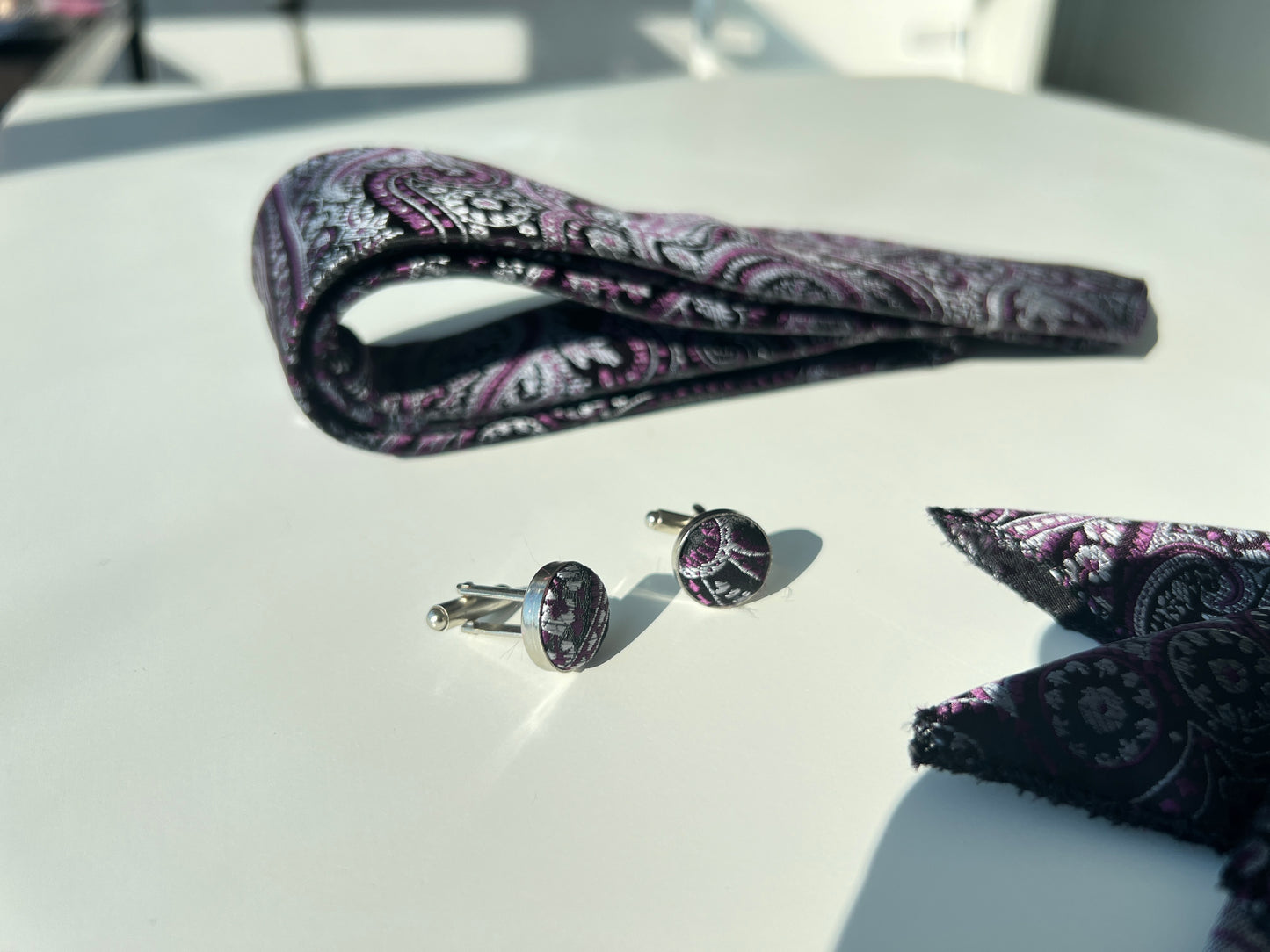 avish purple paisley tie and pocket square set, accompanied by complementing cufflinks, all laid out on a white surface with sunlight enhancing the texture and colors."