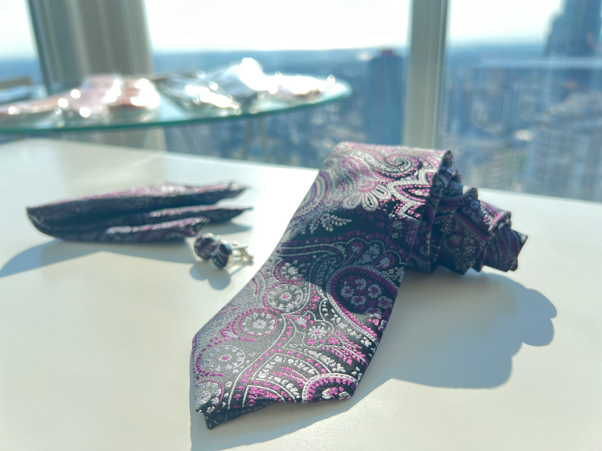 avish purple paisley tie and pocket square set, accompanied by complementing cufflinks, all laid out on a white surface with sunlight enhancing the texture and colors."