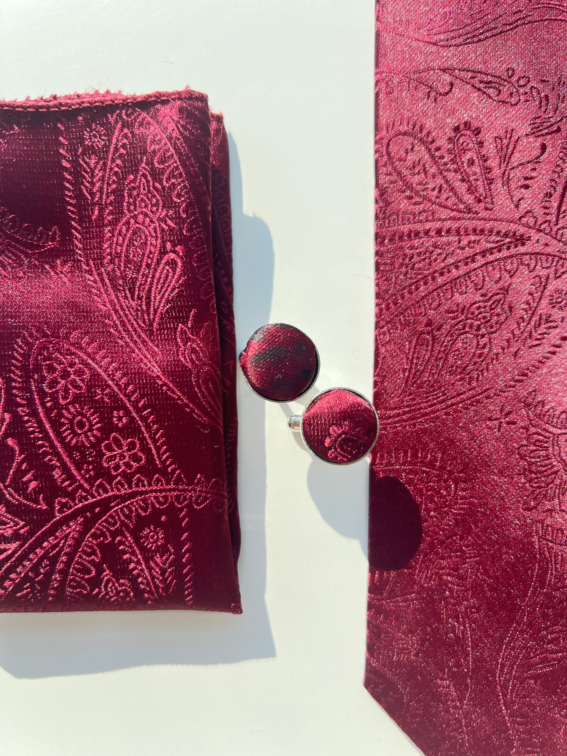 Close-up view of a dark red paisley tie set including a silk pocket square and metallic cufflinks, showcasing detailed embroidery and texture